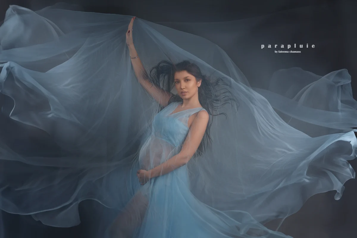 Pregnant woman in flowing blue gown surrounded by sheer fabric, artistic fine art maternity photoshoot at Parapluie Studio Lebanon.