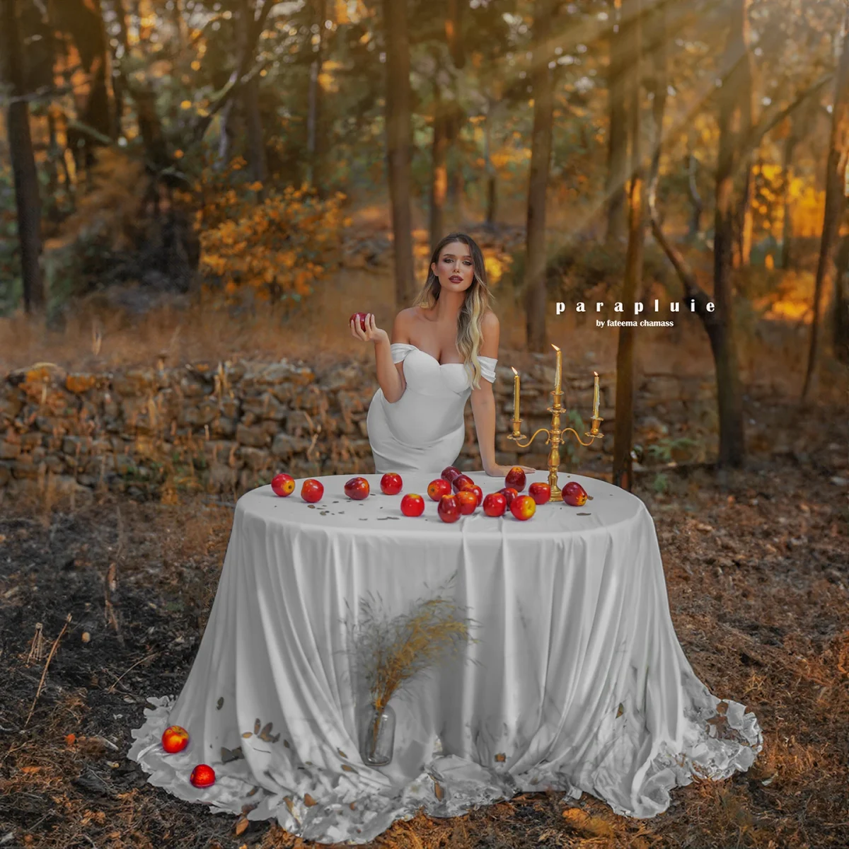 Pregnant woman in elegant white gown standing by a table with apples in a fine art outdoor maternity photoshoot at Parapluie Studio Lebanon.