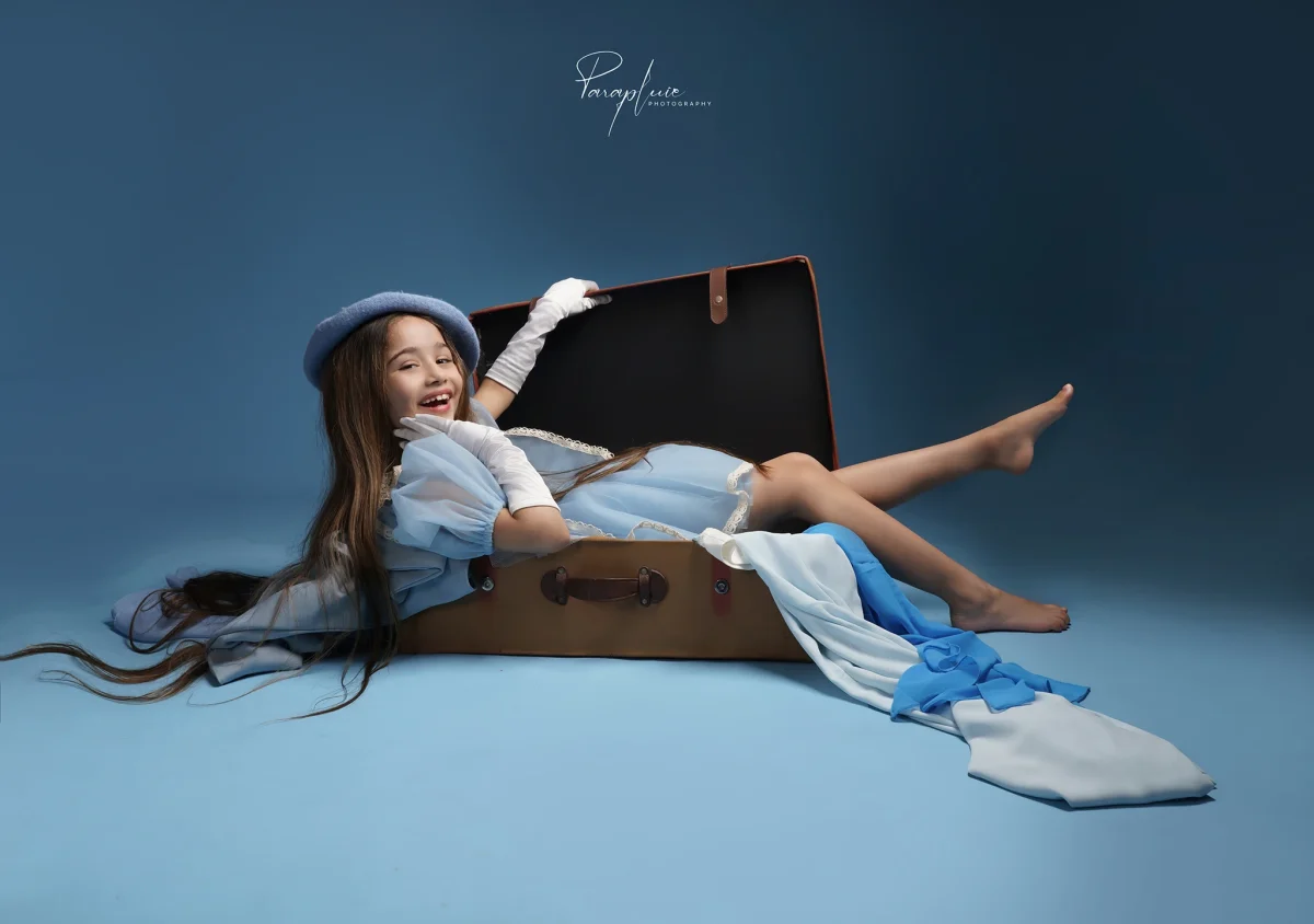 Smiling young girl in a blue dress and hat posing playfully inside an open suitcase, fine art children’s photoshoot at Parapluie Studio Lebanon.