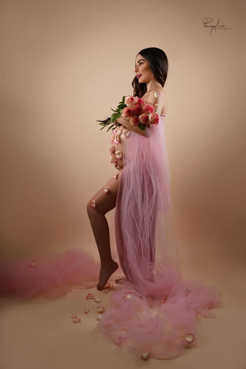 Pregnant woman in pink maternity photoshoot holding a bouquet of roses with scattered petals, fine art pregnancy photography at Parapluie Studio Lebanon.