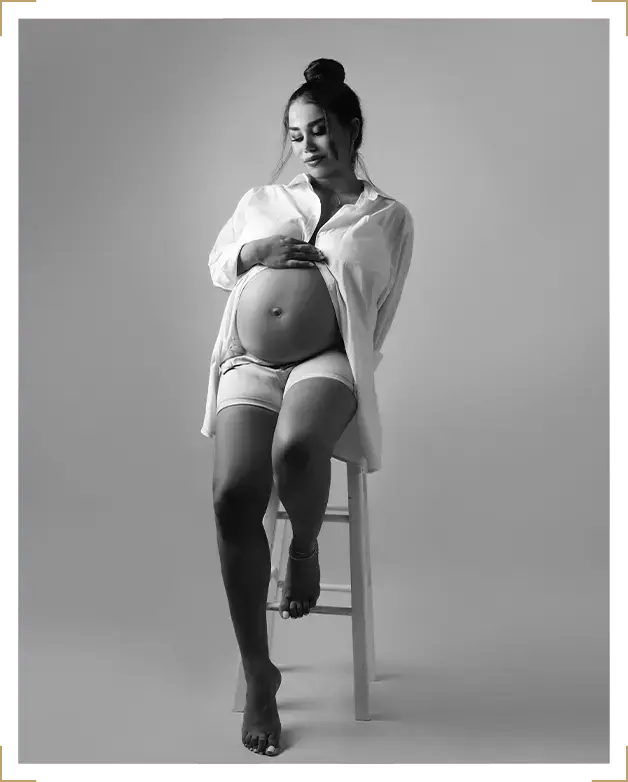Pregnant woman in black and white maternity photoshoot seated on a stool wearing an open shirt and shorts, Parapluie Studio Lebanon.