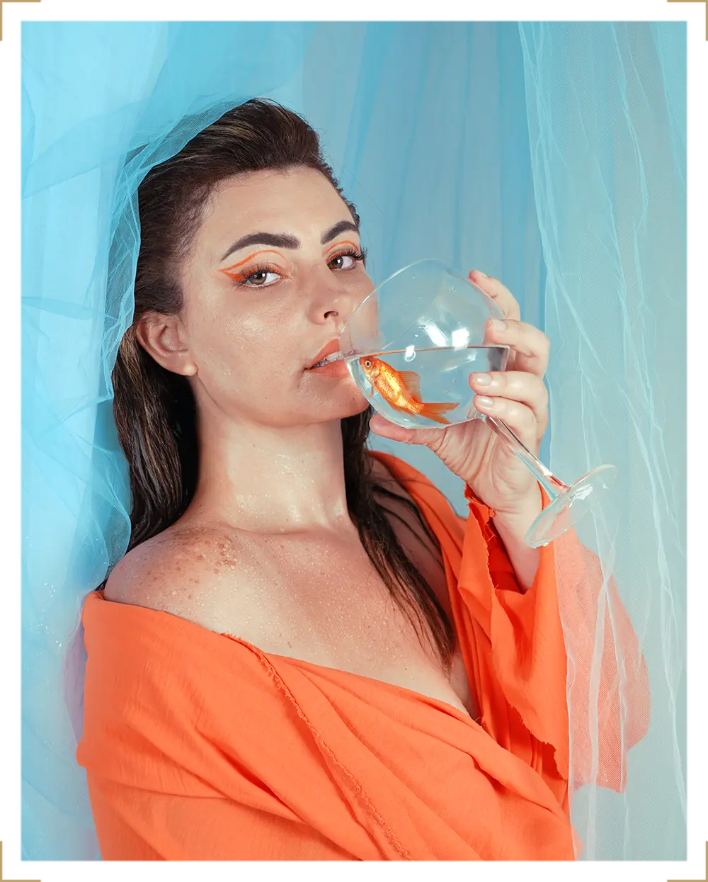 Woman in orange outfit with bold eye makeup holding a glass of water with a goldfish, artistic fashion portrait at Parapluie Studio Lebanon.